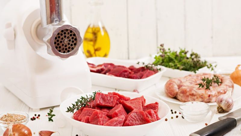 Мясорубка белого цвета, ножи которой иногда нужно затачивать, стоит на столе, а рядом расставлены тарелки с мясными продуктами и зеленью