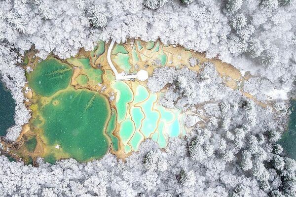 Вид сверху на красочные известняковые бассейны (часто их назвают бассейнами фей) в Национальном парке Цзючжайгоу в провинции Сычуань, Китай. Под нежным снежным покровом бирюзовые и изумрудные узоры в виде драконьей чешуи на дне бассейнов сияют еще ярче.