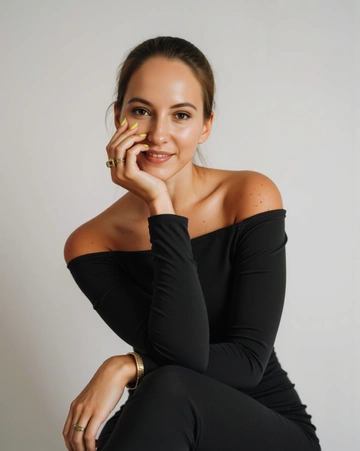 Женщина в чёрном платье с открытыми плечами, улыбается, опираясь щекой на руку.