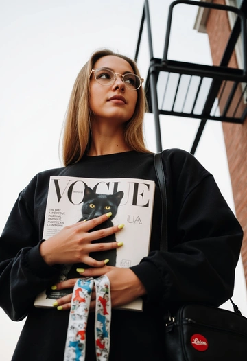 Женщина в чёрном свитшоте с журналом Vogue в руках, позирует на фоне лестницы.