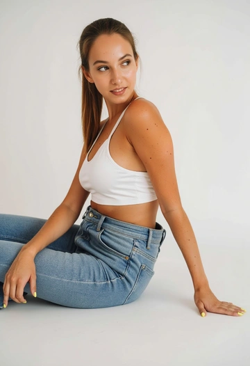 Женщина в белом топе и джинсах сидит на полу в студии с белым фоном.